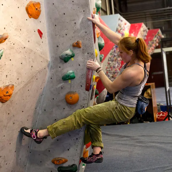 Een vrouw met rood haar beklimt een bouldermuur in een klimhal, waarbij ze zich met zowel haar handen als haar voeten vastklampt aan kleurrijke grepen. Ze draagt een grijze tanktop, een groene broek, klimschoenen en een krijtzak. Op de achtergrond is de k Een vrouw met rood haar beklimt een bouldermuur in een klimhal, waarbij ze zich met zowel haar handen als haar voeten vastklampt aan kleurrijke grepen. Ze draagt een grijze tanktop, een groene broek, klimschoenen en een krijtzak. Op de achtergrond is de k